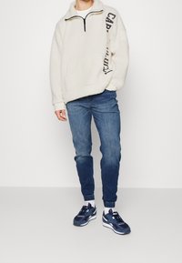 Pullover in pile di colore bianco sporco con zip al collo, con lettering nero. Abbinato a jeans blu scuro e sneaker navy con dettagli bianchi.