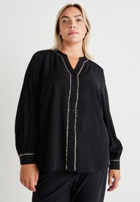 Blusa preta com mangas longas, fecho de botão à frente e tachas prateadas decorativas ao longo da pala e dos punhos. Tecido suave e liso.