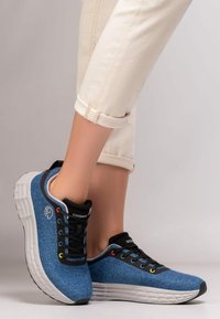 Baskets en tissu bleu avec des lacets noirs, présentant une tige texturée, une semelle blanche et des œillets rouges et jaunes pour un détail d'accent.