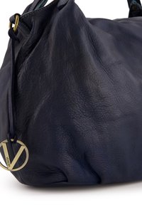 Granatowa skórzana torebka o teksturowanym wykończeniu, z akcentem ze złotego metalu w postaci okrągłego logo oraz miękkim, luźnym kształtem.