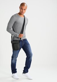 Black messenger bag with adjustable strap, worn across the body; model in a fitted gray long-sleeve shirt and blue jeans, white sneakers.