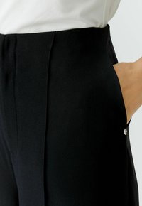 Pantalons noirs avec une texture lisse, dotés d'une poche, d'une fermeture éclair avant discrète et d'un accent de bouton poli près de la taille.
