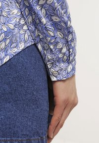 Dlouhý rukáv košile z světlomodré látky s bílým a černým vzorem listů. Má knoflíky na manžetách a dolním lemu. Nositá přes džíny.