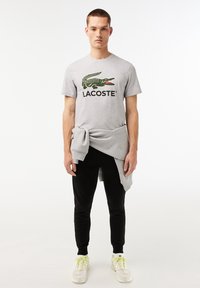 Grå bomullströja med en grön krokodil-logga och svart text "LACOSTE", tillsammans med svarta joggers och vita sneakers med neongula accenter.
