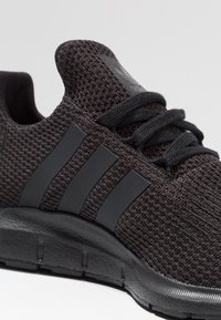 Svart atletisk sneakers med en texturerad mesh-överdel, tre randiga detaljer, platta snören och en dämpad svart sula.