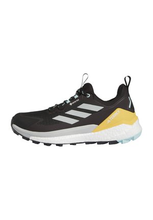 Zwarte trailrunning schoen met een bovenwerk van mesh, zilveren strepen, een gele en witte tussenzool, en een gestructureerde rubberen buitenzool voor grip.
