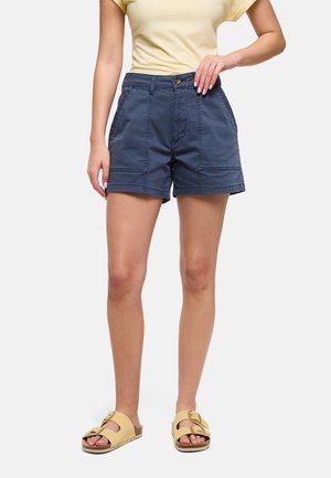 Mujer con pantalones cortos de talle alto azul marino, camiseta amarilla pálida y sandalias beige con hebilla, de pie contra un fondo blanco liso.