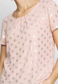 Top rosa claro con un sutil plisado en el escote y lunares metálicos plateados dispersos; mangas cortas y textura de tela suave.