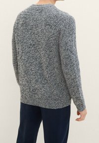 Man in een grijs-wit gemêleerd breiselsweater met lange mouwen en donkerblauwe broek, gezien van achteren tegen een effen achtergrond.