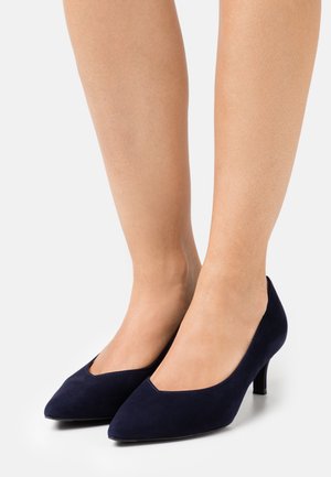 Marineblauwe suède pumps met hoge hak en een spitse neus, voorzien van een v-vormige uitsnede aan de voorkant en een slanke hak van ongeveer 5 centimeter.