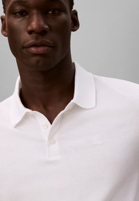 Fehér pamut póló klasszikus gallérral, kétgombos gombolással és diszkrét, a mellkason hímezett CK logóval. Simább textúra.