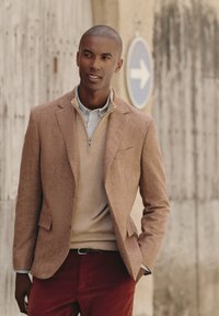 Blazer a spina di pesce marrone sopra un maglione beige, con colletto a righe blu e bianche. Abbinato a pantaloni in velluto a coste rossi.