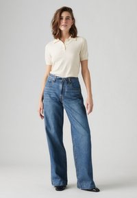 Šviesiai ruda trumpomis rankovėmis polo marškinėliai ir mėlyni aukštu liemeniu platesni džinsai. Lygus audinio tekstūra, kasdienis dizainas ir klasikinis siluetas. Juodi batai.