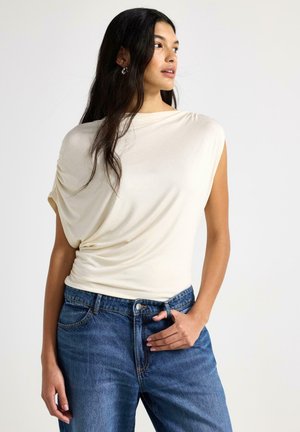 Kortärmad cremefärgad topp i mjukt tyg med draperad design, kombinerad med högmidjade blå denimjeans med framfickor och knappstängning.