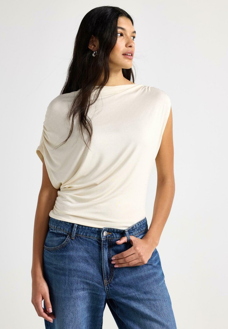 Kremfarvet top med korte ærmer lavet af blødt stof, draperet design, kombineret med højtaljede blå denimjeans med forlommer og knaplukning.