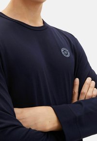 Marinblå långärmad T-shirt i mjukt tyg. Har en rund halsringning och en cirkulär broderad logotyp på bröstet. Plana sömmar.