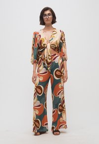 Γυναίκα που φοράει πολύχρωμο σετ με αφηρημένο μοτίβο, αποτελούμενο από μακρυμάνικο τοπ με δέσιμο μπροστά και παντελόνι φαρδιάς γραμμής, στέκεται μπροστά σε απλό φόντο.