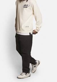 Person wearing a cream sweatshirt with graphic text, black pants, white layered shirt, and white sneakers with black and gray details.
