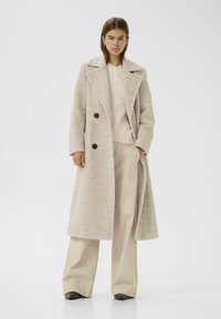 Manteau double boutonnage gris clair avec de grands boutons noirs, manches longues et poches latérales, associé à un pull crème et un pantalon beige à jambes larges.