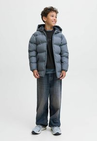 Chaqueta puffer gris con capucha, cierre delantero y puños elásticos; llevada sobre un sudadera negra y combinada con jeans de mezclilla holgados y zapatillas.