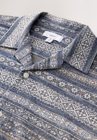 Blaues und beiges Hemd mit kurzem Ärmel und Kragen, mit geometrischen Mustern und Knopfverschluss. Etikett innen am Kragen sichtbar.