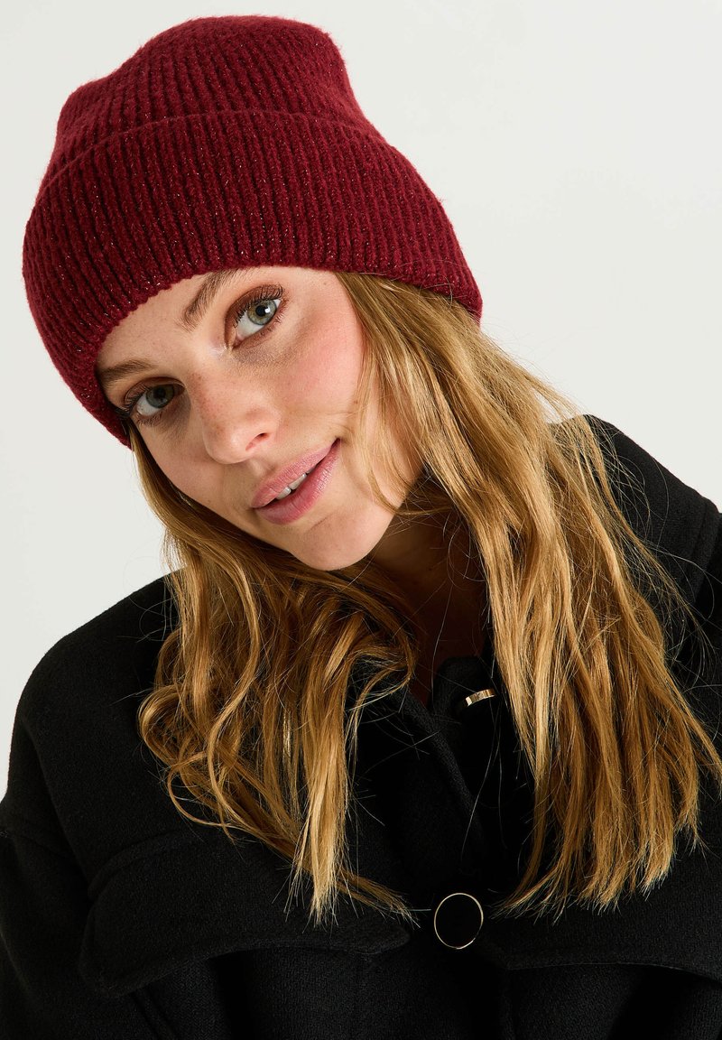 Gorro de punto rojo con textura acanalada. Presenta un ajuste ceñido y un diseño ligeramente holgado. Se lleva con un abrigo negro, resaltando el contraste.