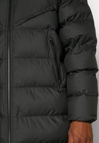 Crna jakna od puf materijala s kvadratnim dizajnom, prednjim zatvaračem i bočnim džepovima sa zatvaračem. Materijal izgleda glatko i izolirano.