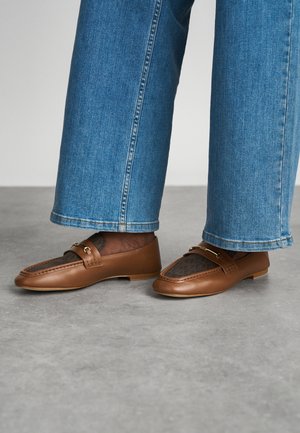 Καφέ δερμάτινα loafers με υφή από καφέ μοτίβο πλέγματος και λεπτομέρειες από χρυσό τόνο, συνδυασμένα με ανοιχτό μπλέ παντελόνια wide-leg.