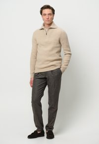 Jersey de punto acanalado beige con cuello con cremallera, combinado con pantalones grises texturizados y zapatos oscuros sin cordones. Fondo liso.