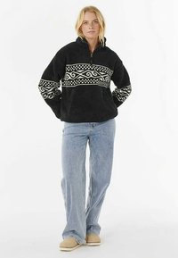 Sudadera polar negra con canesú blanco estampado. Cuenta con cremallera frontal de cuarto de largo, mangas largas y un corte holgado. Combinada con jeans azules.