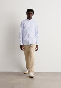 Lys blå knappeløs skjorte med lange ermer, beige bukser og hvite sneakers. Enkelt design, subtil tekstur og tettsittende silhuett.