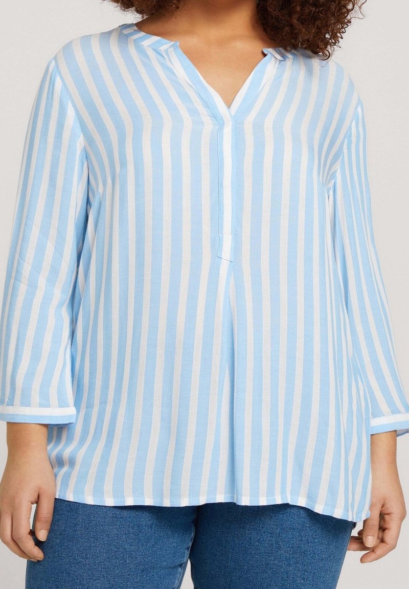 Frau trägt eine hellblau-weiß vertikal gestreifte Bluse mit V-Ausschnitt und dreiviertel Ärmeln.