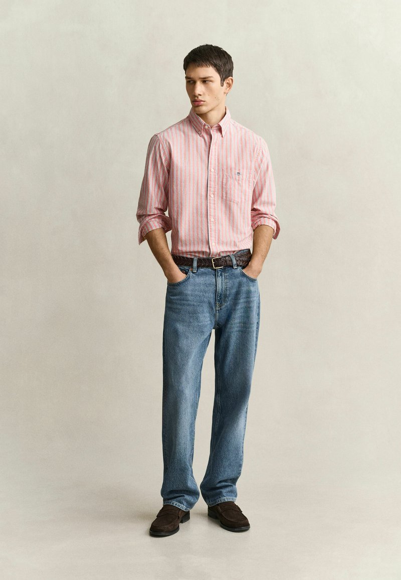 Jeune homme debout, mains dans les poches, portant une chemise à rayures roses et blanches, un jean bleu, une ceinture marron et des chaussures marron foncé sur un fond uni.