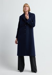 Marineblauer Wollmantel mit Kragen, fünf Knöpfen und seitlichen Taschen, kombiniert mit schwarzen Nadelstreifen-Hosen und hohen Schuhen.