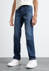 Jean en denim bleu avec un design à jambe droite, présentant des coutures discrètes, des poches avant, et accompagnés de baskets blanches.