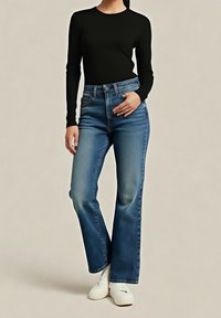 Femme portant un haut noir à manches longues, un jean bleu taille haute et des chaussures blanches, debout devant un fond uni.
