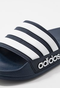 Blauwe slipper met drie witte strepen en zwarte "adidas"-tekst. Heeft een gestructureerd materiaal en een gevoerde binnenzool.