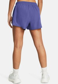 Lila idrottsshorts med elastisk midja. Slätt tyg, lös passform, och shortsen sträcker sig något till övre låret. Ett par vita sneakers synliga.