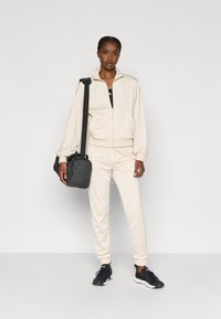 Krämfärgad träningsdräkt av mjukt tyg, med en zipjacka med ståkrage och matchande joggingbyxor. Svarta sportskor och en mörk väska.