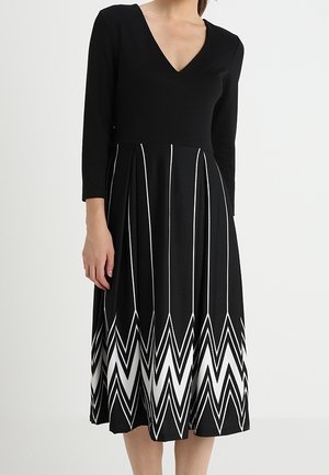 Mujer con un vestido negro de manga larga con cuello en V y patrones de zigzag blancos en la falda plisada.