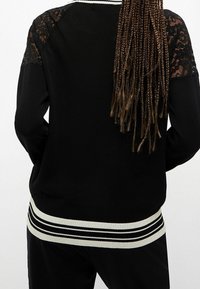 Czarny sweter z koronkowym detalem na ramionach, prążkowym białym i czarnym paskiem na dole, luźny krój oraz miękka, teksturowana dzianina.