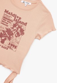 Camiseta de manga corta en color melocotón con un gráfico floral, texto que dice "Isla Maldivas" y un dobladillo sin terminar con un acento atado a un lado.