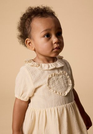 Niña pequeña con cabello rizado que lleva un vestido amarillo pálido con un detalle de corazón con volantes, mirando hacia un lado contra un fondo beige.