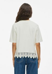 Camiseta de manga corta color crema con un dobladillo de encaje en forma de ola, con una textura de tela suave y un corte relajado. Combinada con unos vaqueros de denim azul.