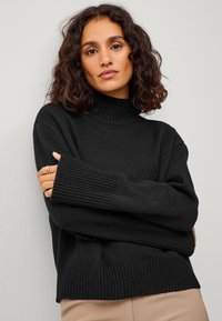 Schwarzer Strick-Pullover mit Rollkragen, gerippten Bündchen und Saum, locker geschnitten und mit texturiertem Oberflächenmaterial. Kombiniert mit beigen Hosen.