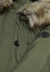 Olivgrön parkas med fuskpälsdetalj, stora svarta knappar och en rund tygpatch på axeln. Slät, texturerad tyg.