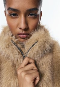 Műszőrme kabát világosbarna színben, puha, texturált gallérral és ezüst cipzár díszítéssel. Kéz, amely a cipzár húzóját fogja.