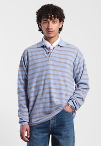 Lichtblauwe polo met lange mouwen en bruine horizontale strepen, kraag en drieknopenlijst. Gekozen in combinatie met blauwe jeans, gemaakt van een gestructureerde stof.