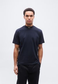 Μπλε ναυτικού τύπου μπλουζάκι με λαιμόκοψη, κατασκευασμένο από βαμβάκι, με χρυσό λογότυπο στο γιακά, κοντά μανίκια και χαλαρή εφαρμογή.