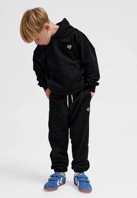 Svart hoodie och mjukisbyxor i mjukt tyg, med en liten logga på bröstet och sidan. Matchas med blå och vita sneakers.